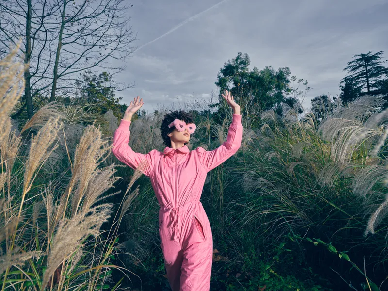 Woman in a pink jumpsuit dancing outdoors in a field of tall grasses, wearing floral sunglasses, expressive pose, carefree and joyful, bright daylight in a natural setting, vibrant fashion moment