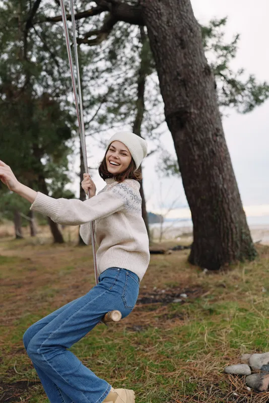 Woman on a swing in a park, wearing a knit sweater and beanie, smiling as she enjoys an outdoor moment among trees and grass.