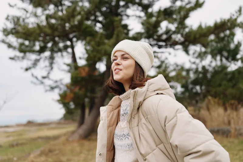 Woman outdoors wearing a beige puffer jacket and knit beanie, standing in a windy natural landscape with trees in the background, looking up with a calm expression.