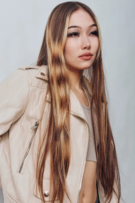 Young woman with long straight hair wearing beige jacket and top standing against neutral background looking aside with calm expression and natural makeup.