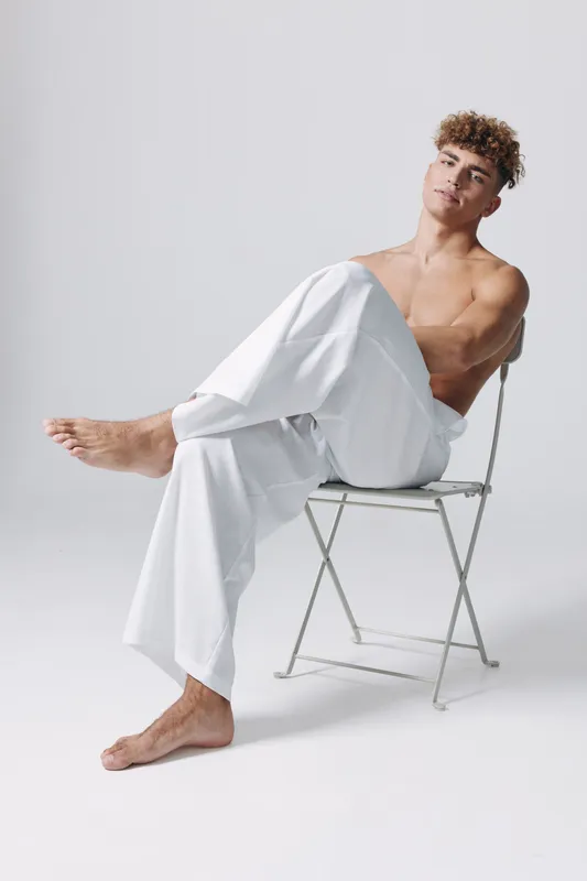 Young man with curly hair, shirtless and wearing white pants, sitting relaxed on a metal chair in studio with clean white background, barefoot and casual posture.
