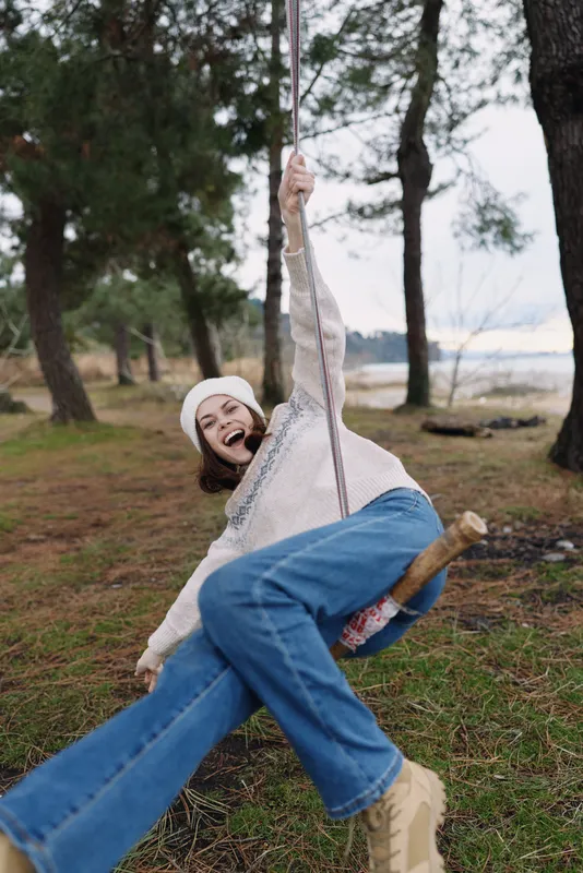 Joyful woman swinging outdoors in a forest park, laughing and leaning back on a rope seat, wearing a beanie, sweater and jeans. Vivid trees and grass create a carefree, candid moment of play.