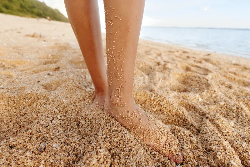 Close up of bare feet sinking into soft beach sand, with the sea and sky in the background. A calm moment illustrating relaxation, summer and grounding by the shore.