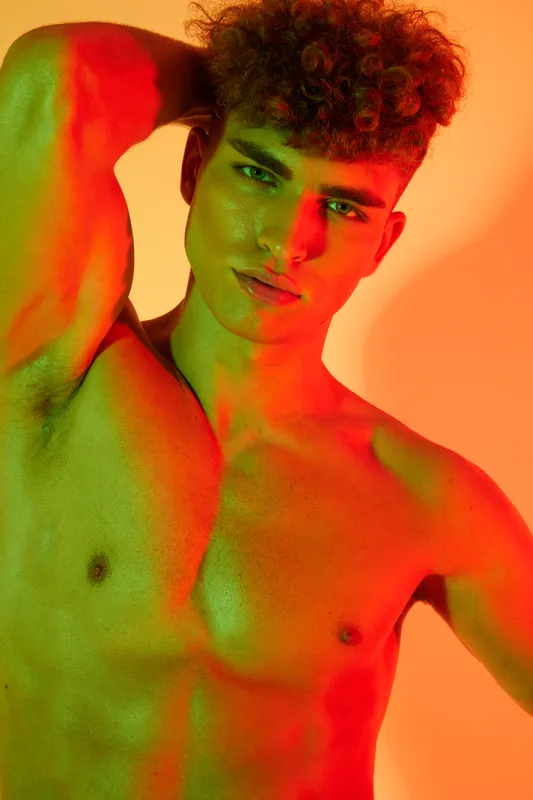 Young man with curly hair posing shirtless with hand behind head under colorful red and green lighting for fashion portrait and artistic expression in studio.