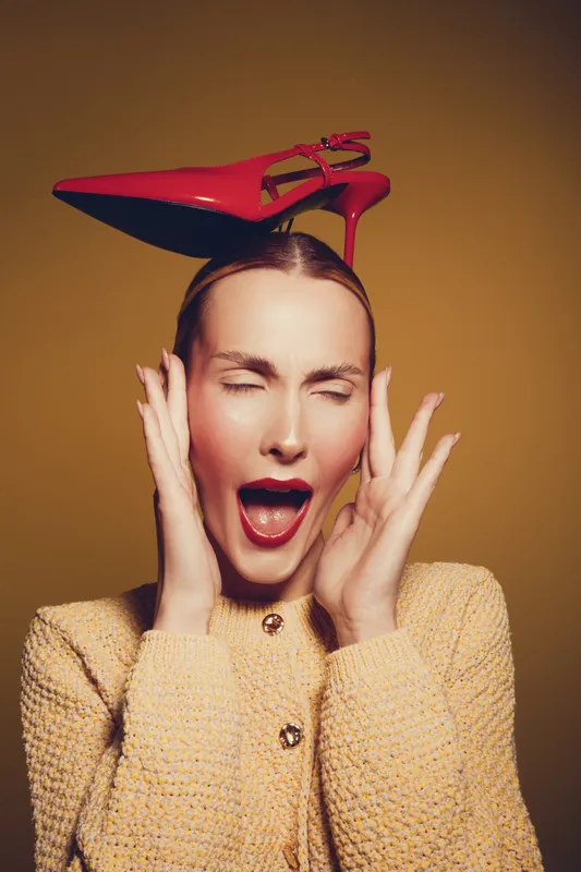 Young woman with red high heel shoe on her head expresses surprise with closed eyes and open mouth. Studio portrait on brown background. Concept of fashion and unusual style.