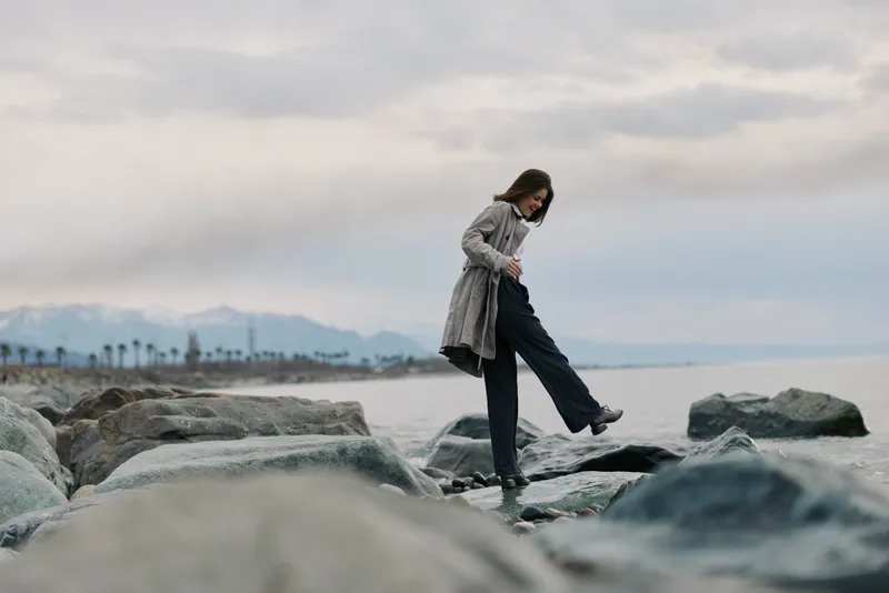 Woman in grey coat and black pants carefully steps on rocks near calm sea under cloudy sky. Concept of balance, nature, outdoor activity, and cautious movement by water.