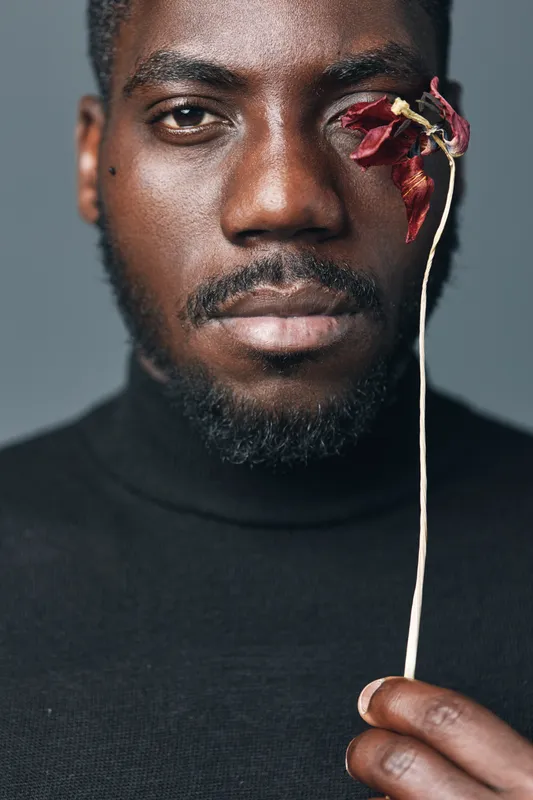 African man with dark skin holding dried red flower near face in creative conceptual visual style. Closeup portrait with serious expression and black turtleneck on gray background.