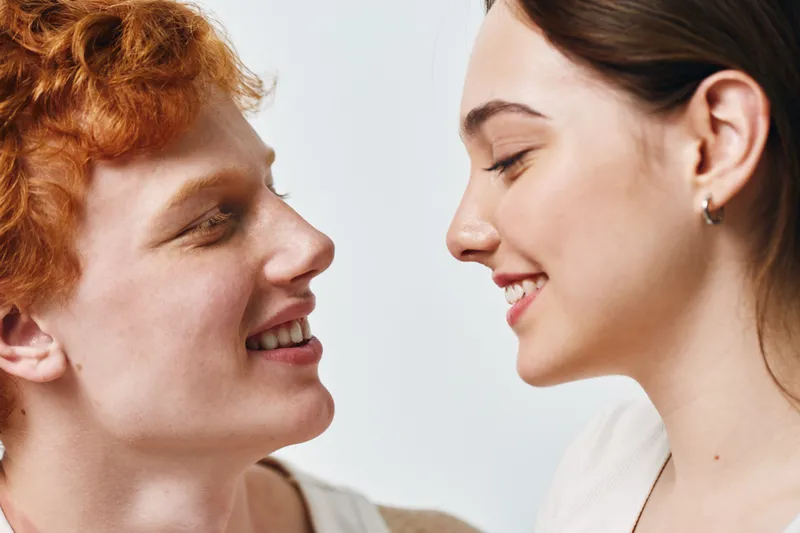 Young man and woman smiling and looking at each other closeup with happiness and affection in studio portrait with bright background and natural expressions.