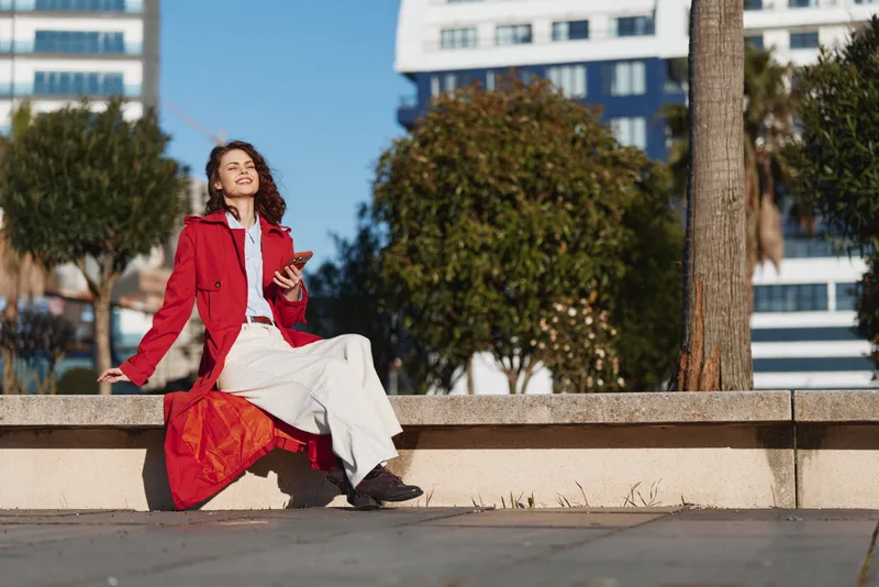 Young woman in red coat sitting outdoors on concrete ledge using smartphone sunny day casual outfit relaxed mood urban background