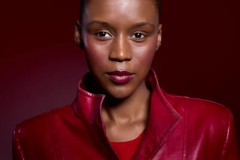 Close-up portrait of a confident African woman with dark skin wearing a red leather jacket on dark red background. Concept of fashion, beauty, and empowerment in studio lighting.
