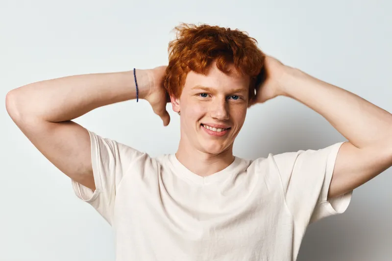 Young man with red hair and freckles wearing a white t shirt smiling with hands behind head on white background. Casual confident male posing relaxed and happy in studio.