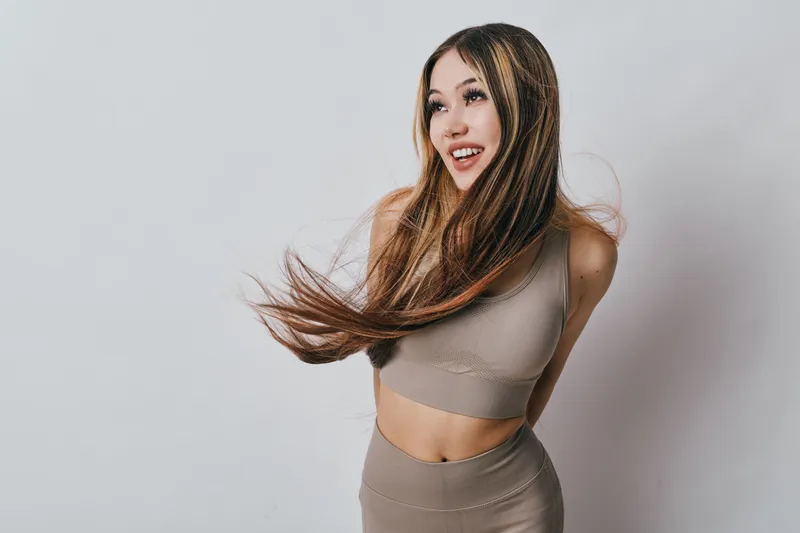 Young woman with long hair wearing beige fitness outfit posing smiling with movement against white studio background, healthy lifestyle and casual confident look.