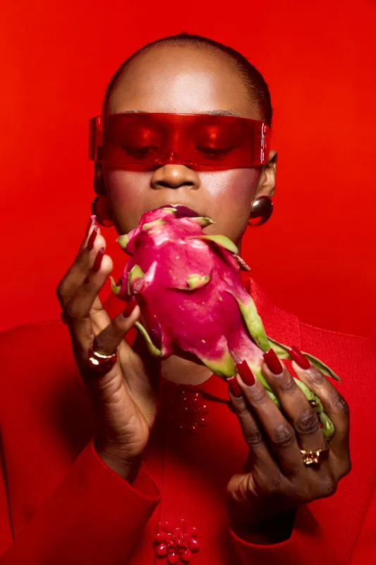 Dark skinned African woman holds dragon fruit close to face with closed eyes wearing red outfit and translucent red sunglasses on red background. Concept of exotic fruit and fashion.