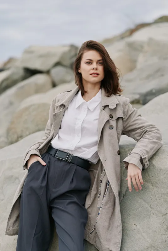 Confident young woman in stylish coat and white shirt poses outdoors on large rocks. Fashion portrait with natural light and casual elegance. Concept for autumn style and modern look.