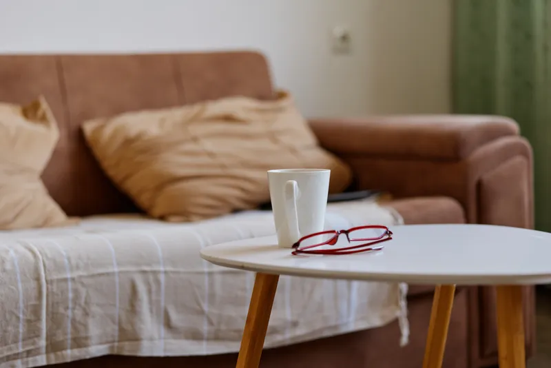 Cozy living room scene with a round table, cup and glasses, soft natural light, comfortable sofa, cushions, warm tones, calm home atmosphere and inviting daytime stillness