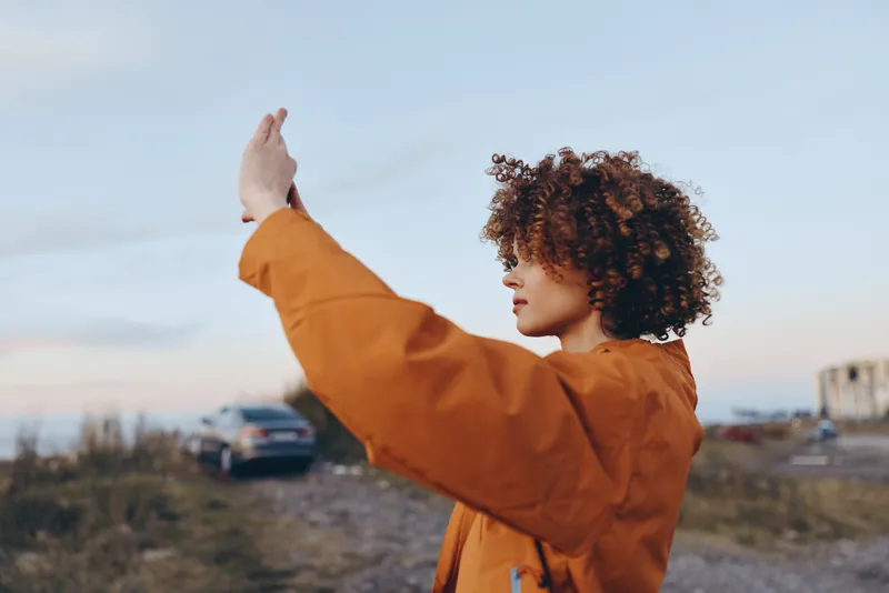 Woman lifestyle outdoors, curly hair, orange coat, arms raised at sunset on road, urban horizon behind, serene calm mood, soft bokeh, natural light, lifestyle portrait