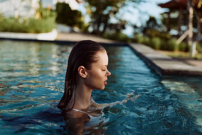 Woman in a pool, profile visible above water, enjoying tranquil outdoor leisure moment, sunlit ripples around her, calm atmosphere, lifestyle scene focused on personal refreshment.