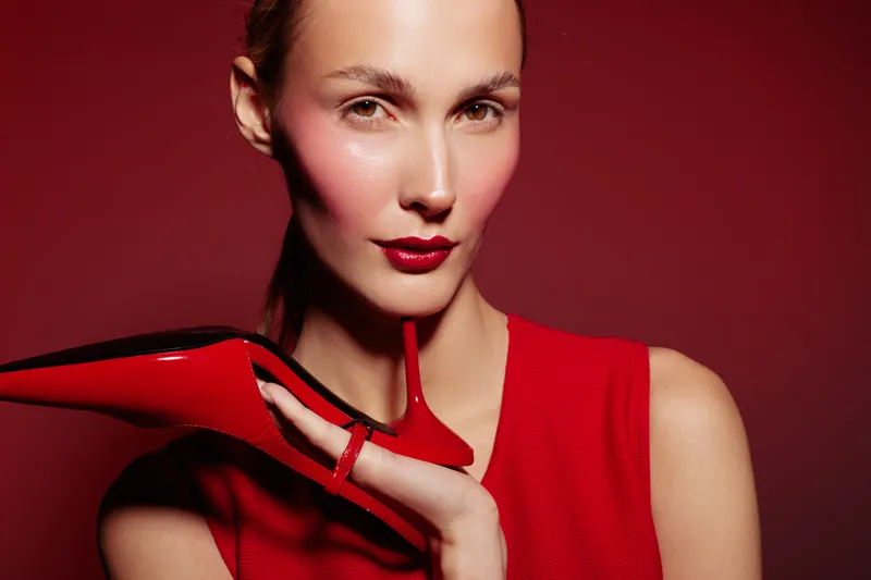 Close-up of a stylish woman in red dress holding a red high heel shoe near her face on red background. Concept of fashion, beauty, elegance, and glamour with vibrant colors.
