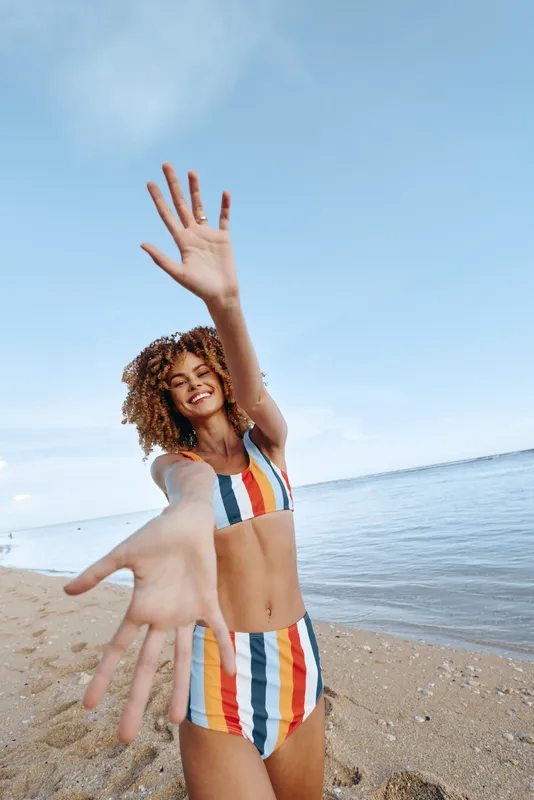 Young woman in a striped swimsuit playfully reaches toward the camera on a sunlit beach, smiling with a relaxed pose as the sea and clear sky form a vibrant background