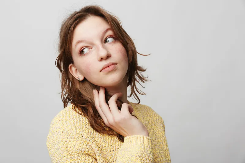 Young woman in yellow sweater looks sideways with thoughtful expression, touching her neck. Studio portrait on grey background. Concept of contemplation, emotion, and casual fashion.