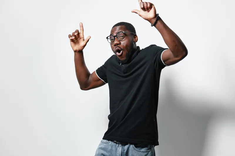 African American man with dark skin wearing glasses and black t shirt posing with excitement and raised hands on white background. Casual style and joyful expression.