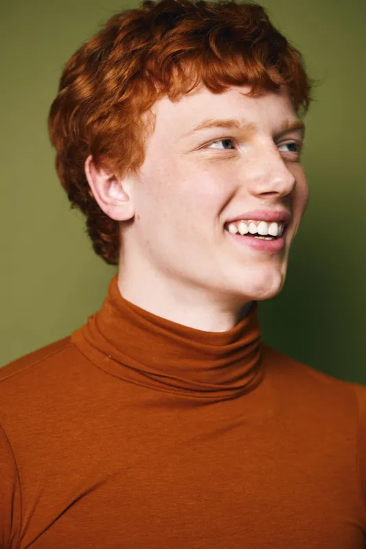 Portrait of a redhead young man smiling and looking sideways wearing a brown turtleneck against a green background with natural light and cheerful expression.