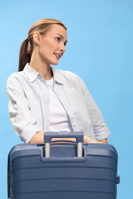 Smiling woman with ponytail holds blue suitcase on light blue background. Travel concept with casual clothing and positive mood, ready for vacation or business trip.