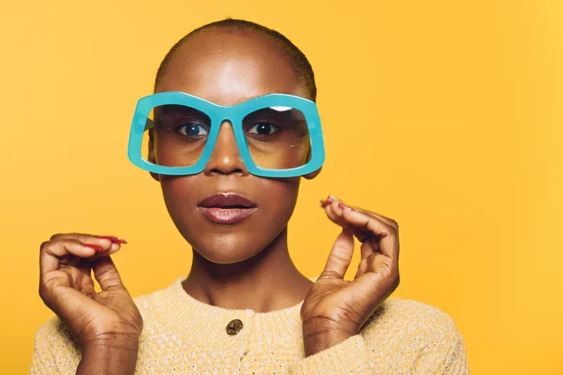 Close-up of confident African woman with dark skin wearing oversized blue sunglasses and beige sweater against yellow background. Concept of fashion, style, and beauty portrait.