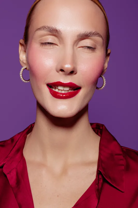 Close-up of a smiling woman with red lipstick and hoop earrings wearing a red blouse on purple background. Concept of beauty, makeup, fashion, and confident expression.