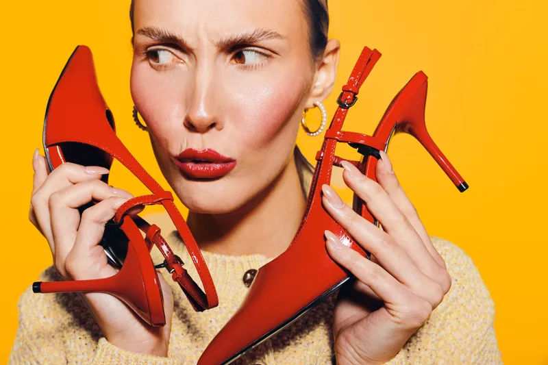 Young woman with red lipstick holds two red high heel shoes near her face, looking confused. Bright orange background highlights fashion and style concept.