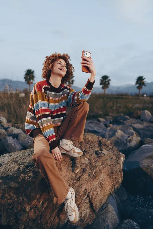 Woman lifestyle smartphone selfie outdoors travel rocks desert, sitting on a boulder in sunny landscape, curly haired woman in colorful sweater and casual pants holds smartphone, smiling for a photo,