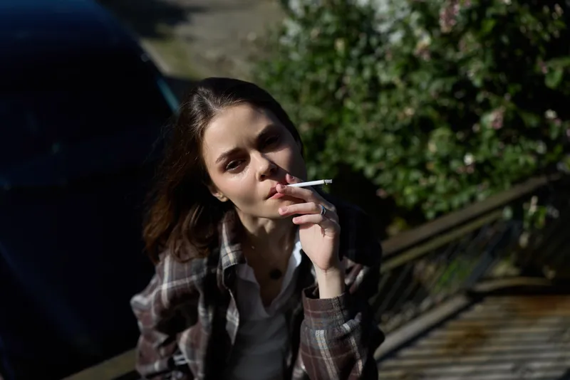 Young woman outdoors leans against a railing, casually smoking a cigarette with a relaxed, contemplative expression, wearing a plaid shirt and sunlit natural surroundings, captured in a candid