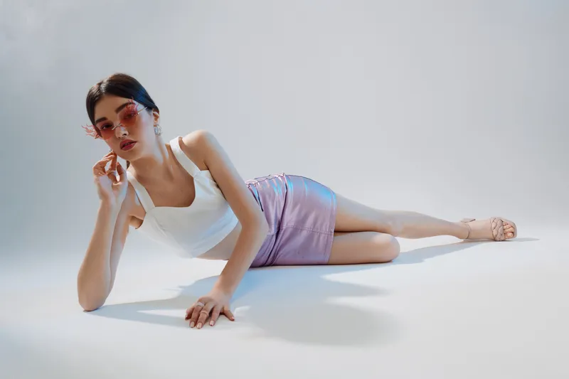 Young woman in white top and purple skirt poses lying on the floor with sunglasses. Studio shot with soft lighting. Concept of woman beauty fashion and style.