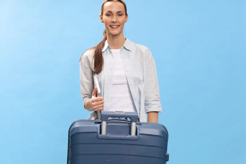 Young woman traveler with suitcase and passport stands against blue background, smiling confidently. Concept of travel, vacation preparation, and tourism for modern lifestyle.