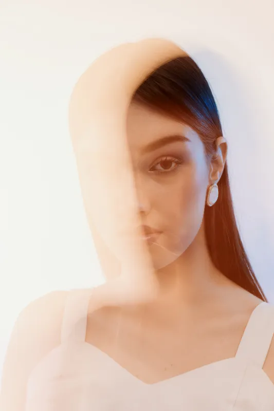 Portrait of a young woman with long hair wearing white earrings and a white top. Soft lighting creates a dreamy fashion and beauty concept with artistic blur effects.