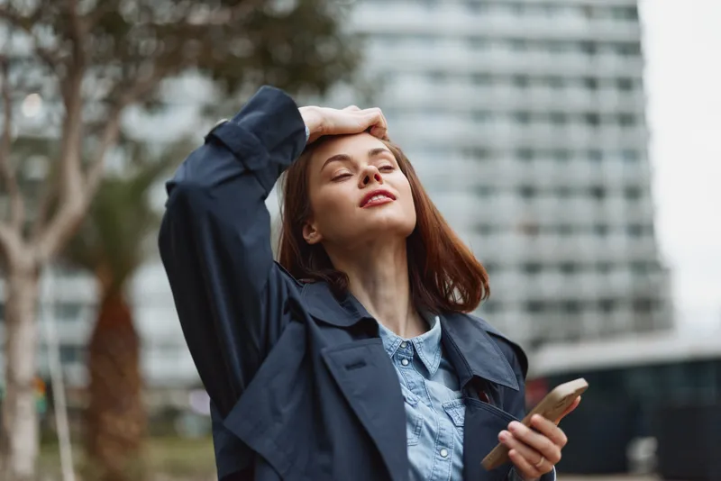Young woman with redhead hair wears a denim jacket and blue shirt, standing outdoors with eyes closed, face lifted to the sun, radiating calm and warmth.