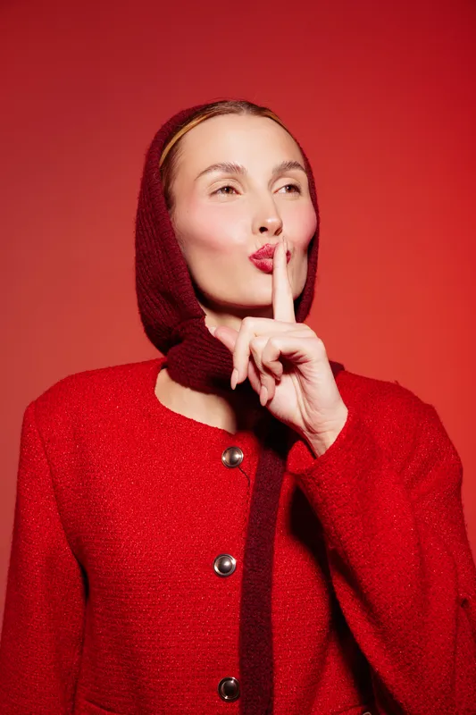 Woman in red coat and knitted hood holds finger to lips in shushing gesture. Studio portrait on red background with soft lighting. Concept of silence, secrecy, and quiet communication.