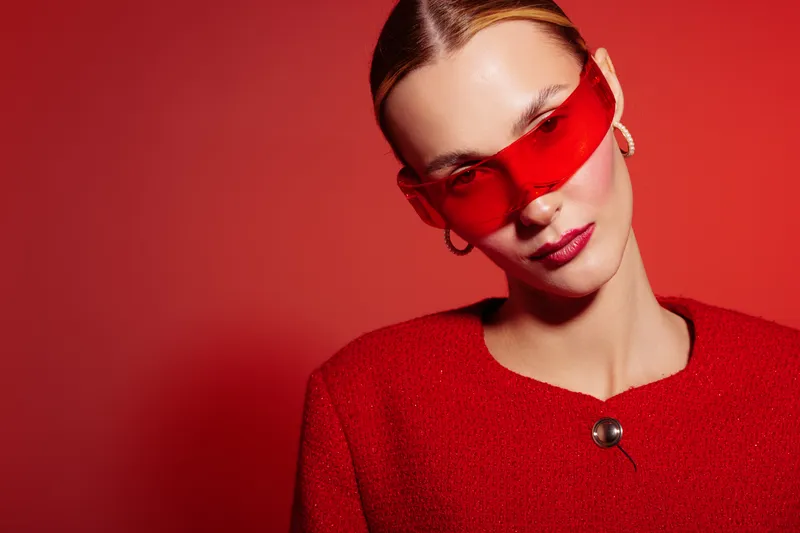 Stylish woman with slicked hair wearing red sunglasses and red jacket poses against red background. Fashion portrait with bold colors and confident expression in studio lighting.