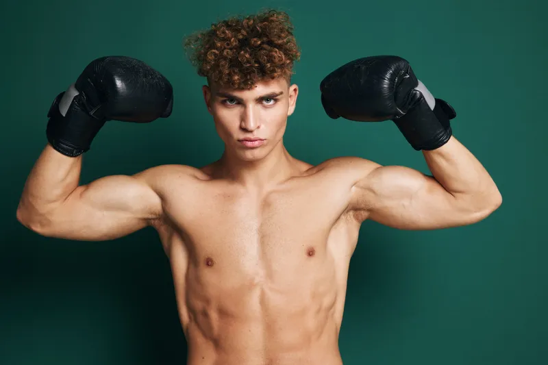 Young muscular man with curly hair wearing black boxing gloves flexing biceps and posing shirtless against green background for fitness and strength concept.