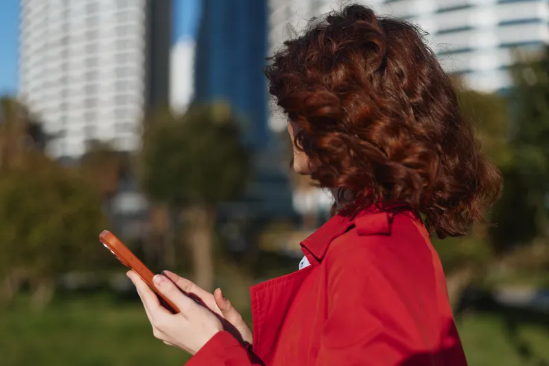Brunette woman in a red coat stands outdoors in an urban park, tapping a smartphone screen with a focused expression against a bright city backdrop today.