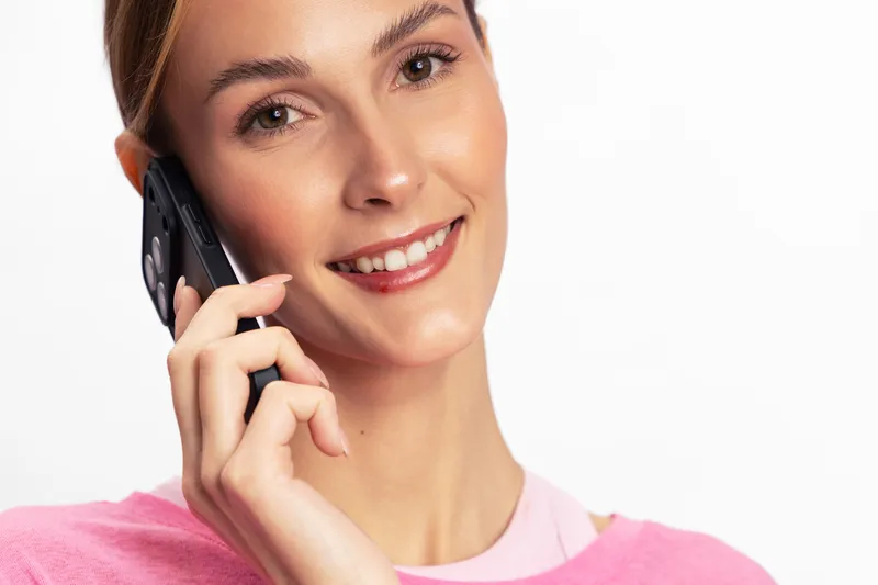 Smiling young woman in pink shirt talking on mobile phone, showing positive emotion and connection. Close-up portrait with white background, concept of communication and happiness.