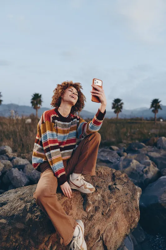 Woman lifestyle outdoors selfie moment, curly hair smiling, striped sweater and tan pants, holding smartphone on rocky path, sunlit palms and distant hills, peaceful pose