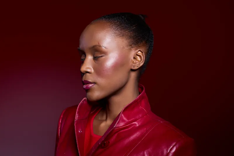 Profile of a calm black woman with dark skin and natural hair wearing a red leather jacket against dark red background. Concept of fashion, beauty, and elegance in studio lighting.