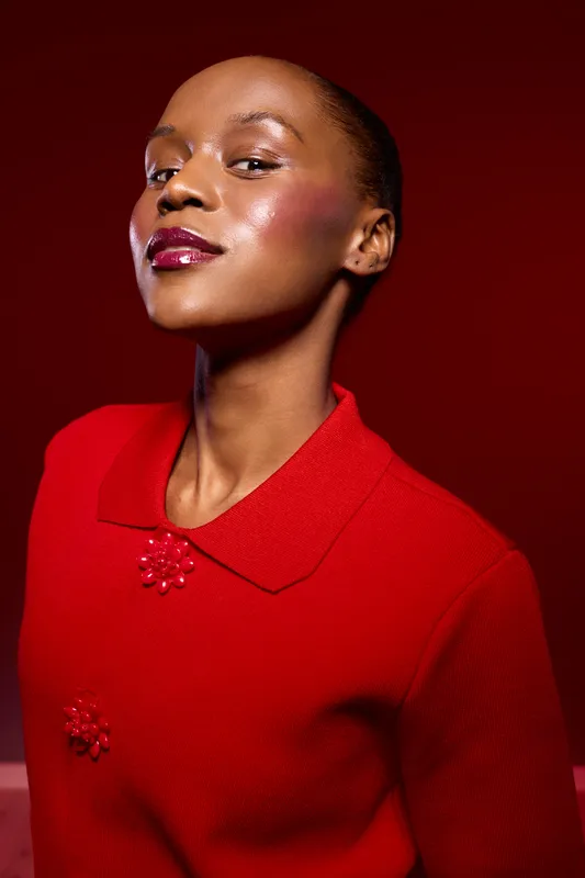 Confident black woman with dark skin wearing a red sweater poses against a red background. Studio portrait of African ethnicity with glossy makeup and elegant expression.
