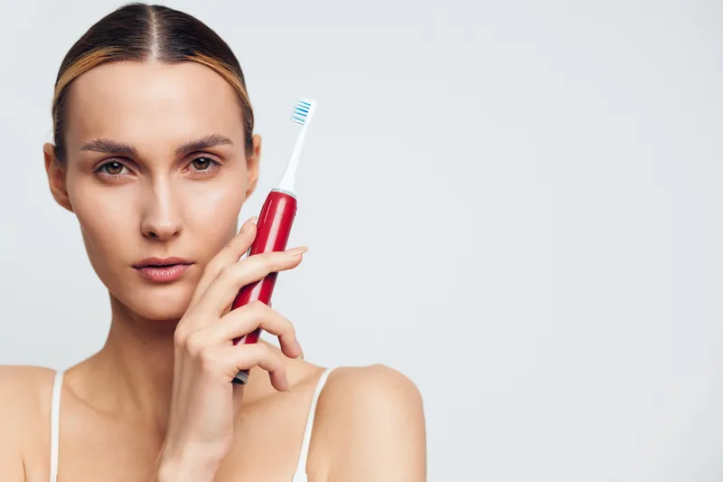 Young woman with slick hair holds a red toothbrush up to her mouth, standing against a plain light backdrop as she concentrates on brushing, conveying a calm, everyday oral care moment.