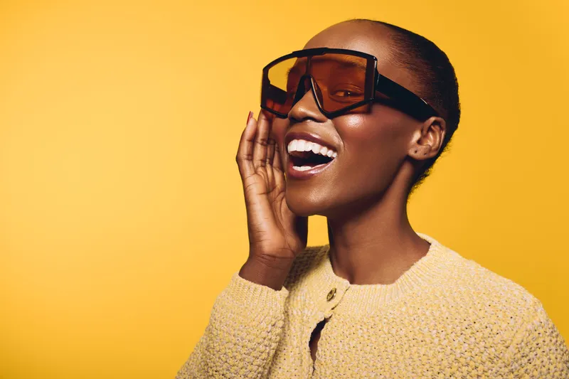 Happy African woman with dark skin wearing large sunglasses and a yellow knitted sweater smiling joyfully on yellow background. Concept of fashion, beauty, and positive mood.