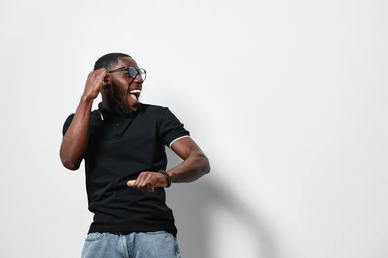 African American man with dark skin wearing glasses and black polo shirt dancing and smiling on white background with casual style and joyful expression.