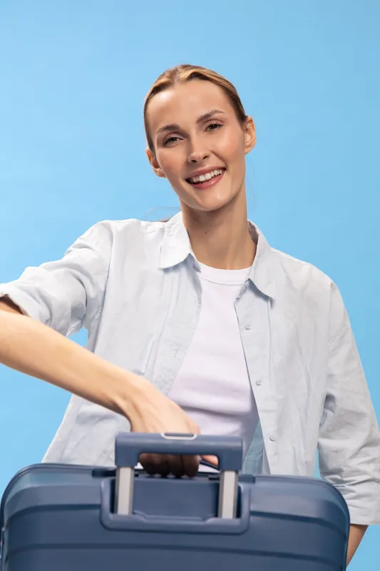Smiling woman with casual shirt holds blue suitcase handle, looking at camera. Travel concept with happy female traveler on blue background, packing or preparing for trip.
