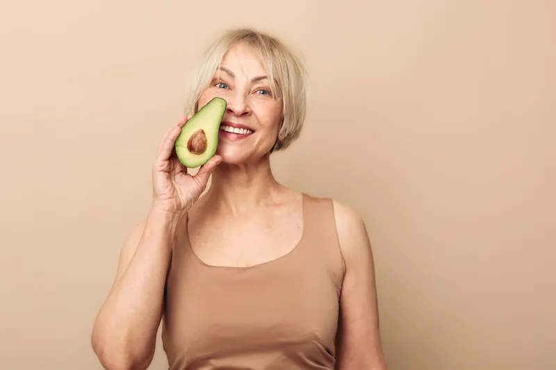 Smiling mature woman holds half of avocado near eye, posing in beige tank top on neutral background. Concept of healthy lifestyle, natural beauty, organic food, wellness and self-care.
