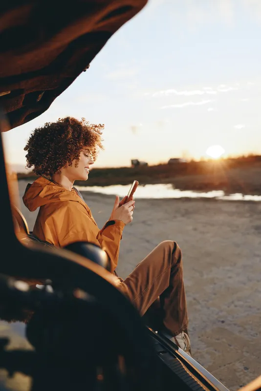 Woman lifestyle at sunset, curly hair visible, young woman sits in a car by the road, holding a drink straw, outdoor golden hour light over ocean water, relaxed travel mood